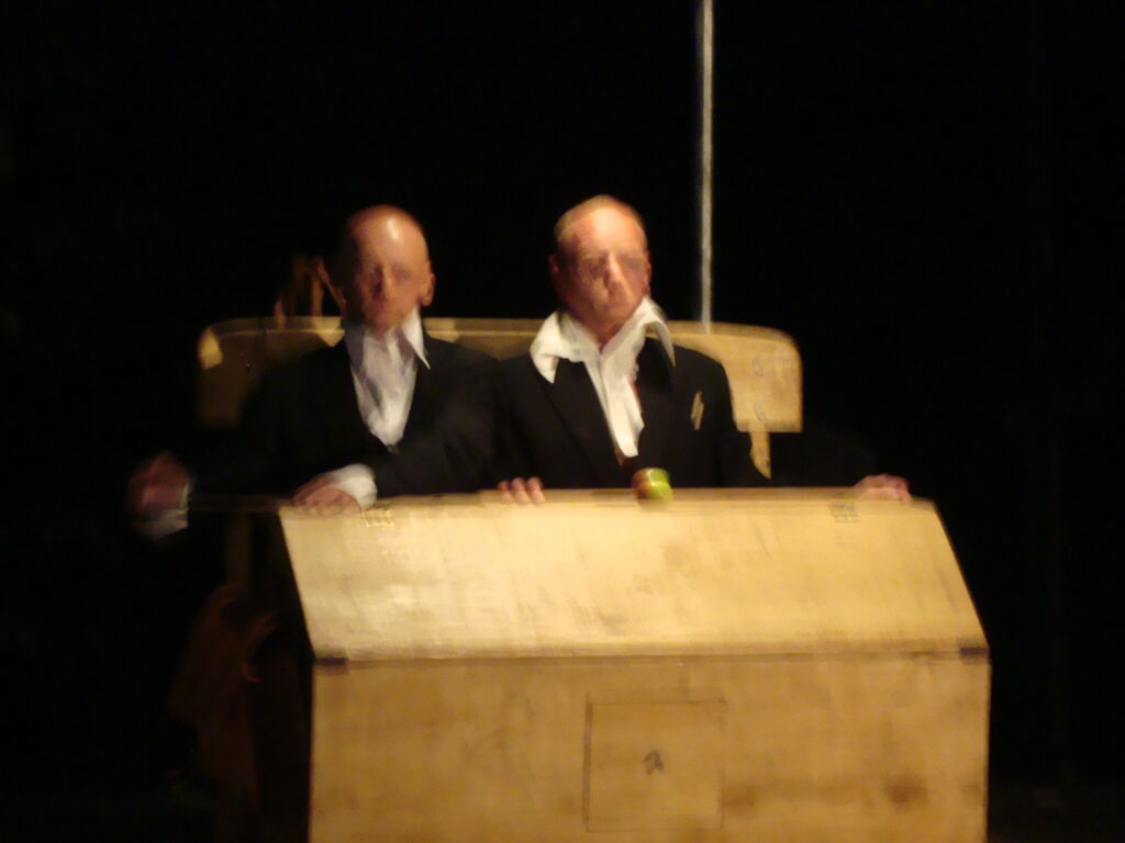 Dwie postacie w strojach, siedzące w drewnianym skrzyni.