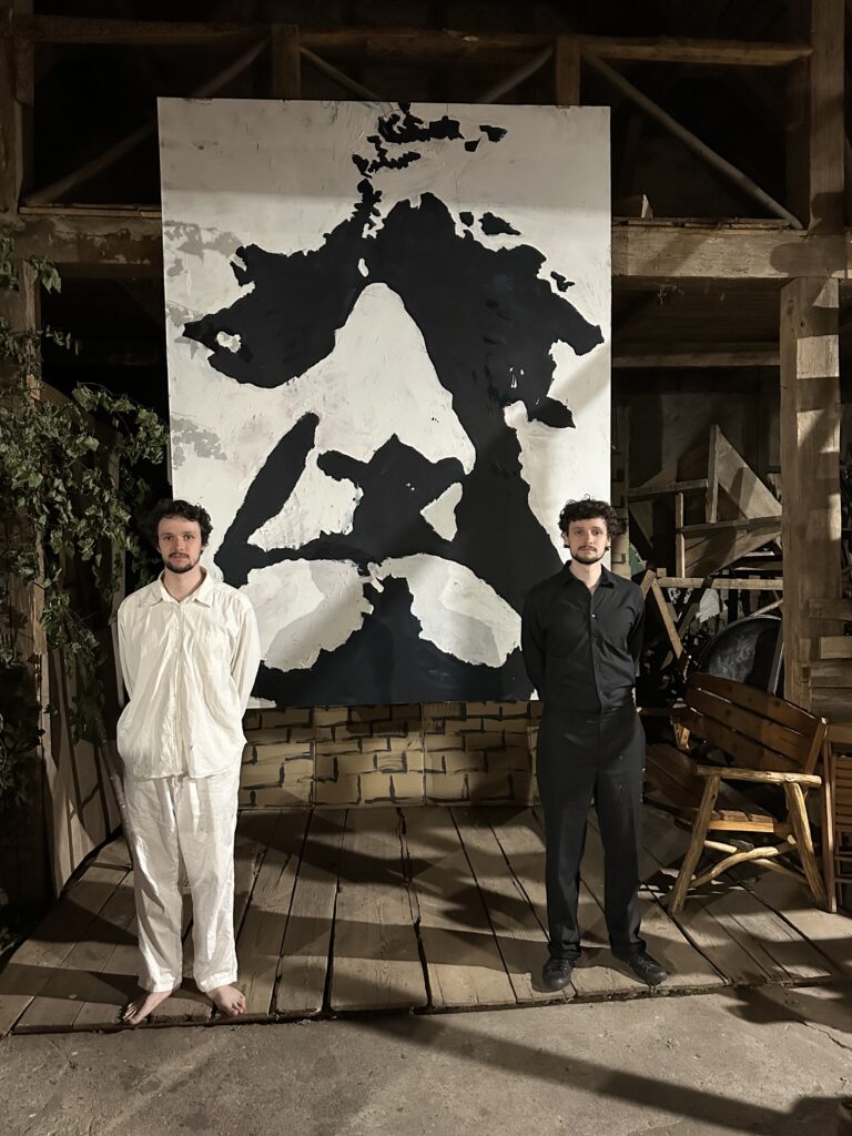 Dwóch mężczyzn przed abstrakcyjnym obrazem na scenie.